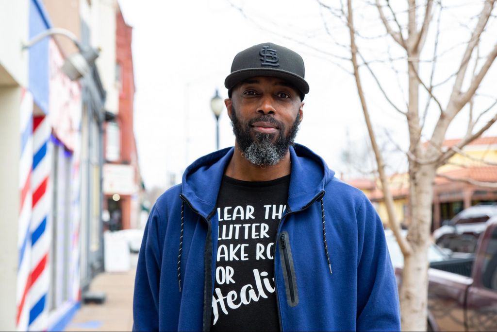 Ronnie Amiyn, a Black man, is facing the viewing with a blue hoodie and a black baseball cap. He has a beard with black and gray hair in it and kind eyes. His t-shirt says, "Clear the clutter or make room for healing."