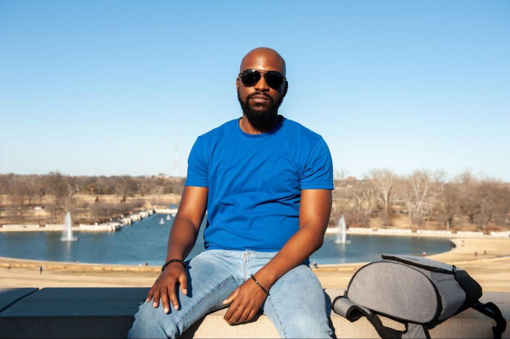 Erik is a Black man is sitting on the ledge in the sun, with the Forest Park Art Hill fountains in the background. It's a bright sunny day and he is bald, wearing sunglasses, has a goatee, and is wearing a blue t-shirt with blue jeans. He has a neutral expression on his face.