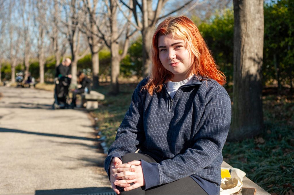 A smiling young white woman with orange and blonde hair and brown eyebrows. She is sitting on a bench in Forest Park with tree trunks in the background and is wearing a zip up long sleeved shirt that is navy with gray cross hatching pattern. She is smiling with a closed mouth and her eyes look kind.