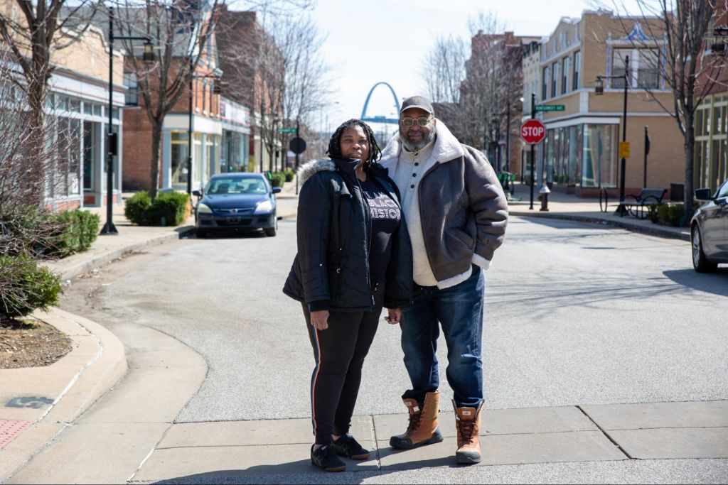 Alison and Riley are a Black couple standing in the middle of the road in North St. Louis City. The St. Louis arch is in the background. Alison has medium length black hair with highlights in twists. She is wearing a puffy coat over a shirt that says, "I am Black history" with long black pants and black shoes. Riley is wearing a baseball cap, has round glasses, and a salt and pepper beard. He is wearing a warm shearling jacket over a knit turtleneck, jeans, and boots. They are look forward with slight smiles.